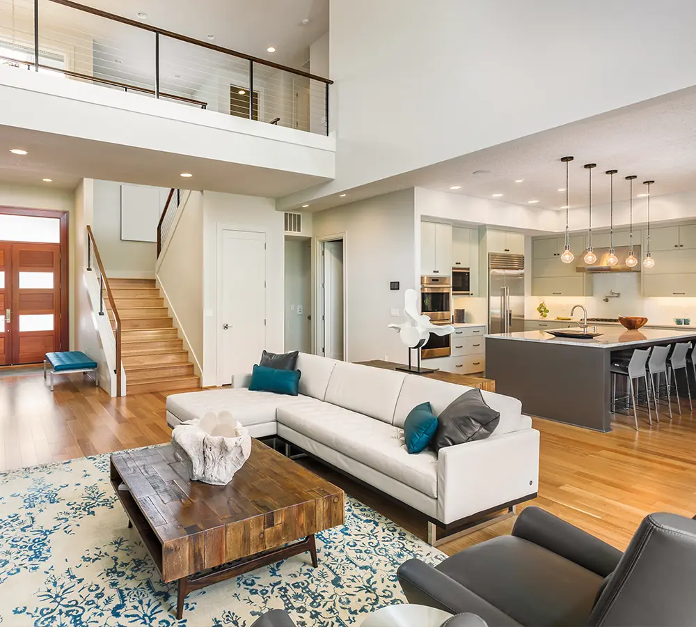 Modern open-concept living room with white sectional sofa, wooden coffee table, and kitchen with pendant lighting.