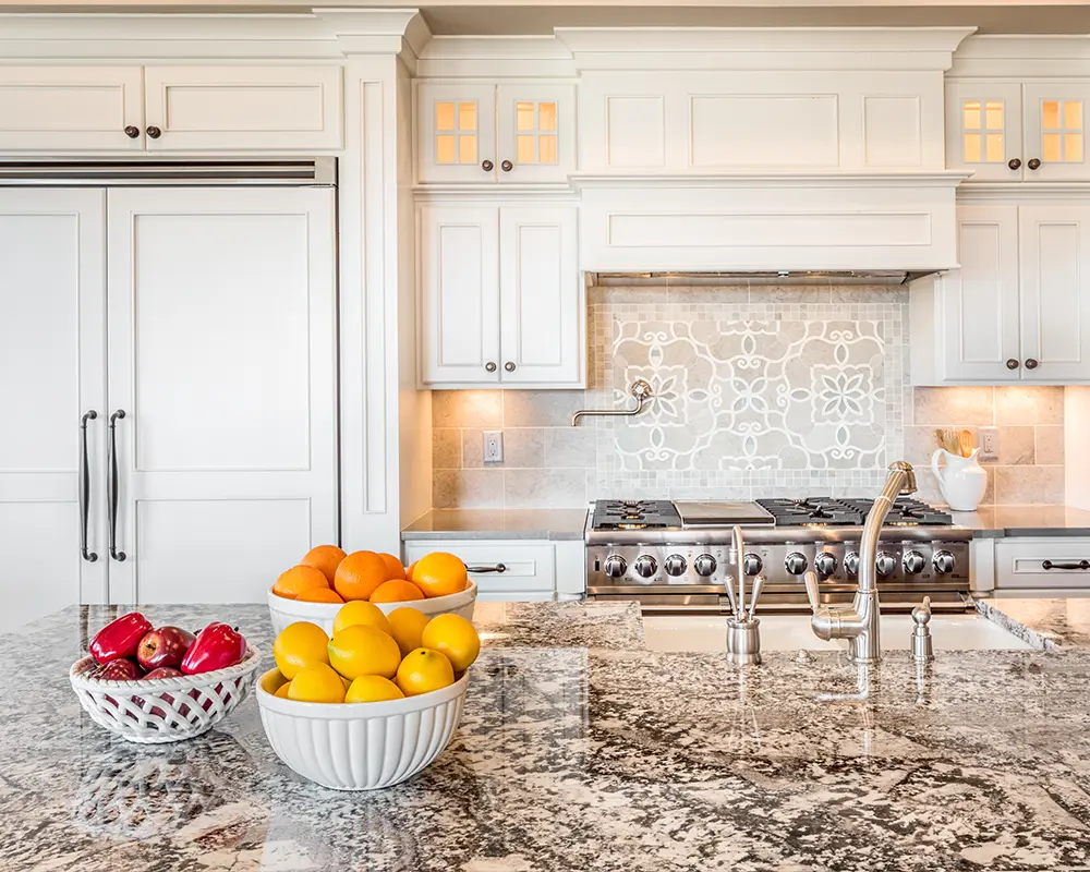 Traditional kitchen showcasing white cabinets, detailed tile backsplash, and fruit bowls on a granite countertop.