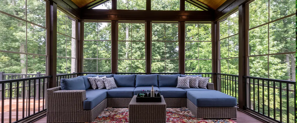 Spacious sunroom with wicker sectional sofa, colorful rug, and vaulted wood ceiling overlooking trees
