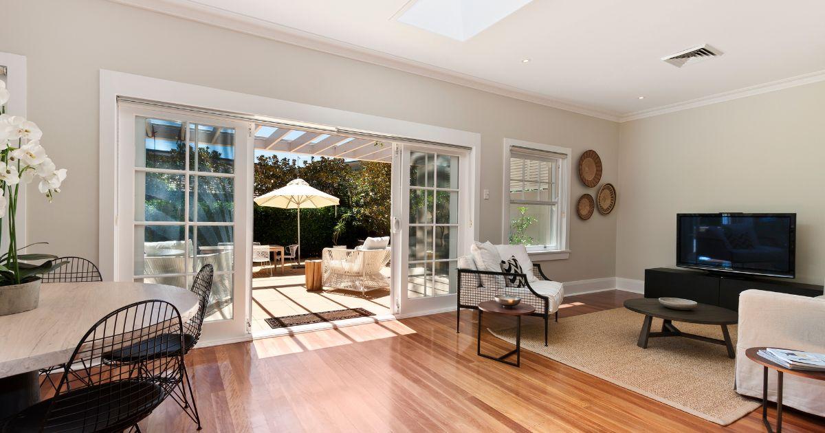 A bright living space with open sliding glass doors leading to an outdoor patio area with seating and umbrellas.
