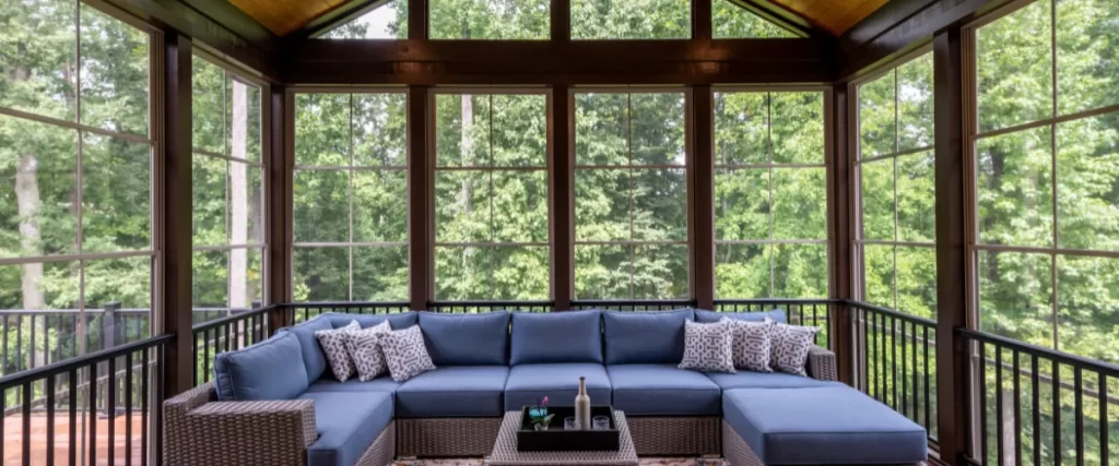 A finished sunroom or enclosed patio with large windows and a blue sectional sofa facing a forested view.